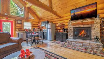 L’intérieur chaleureux d’un chalet en rondins, avec une cheminée en pierre, des poutres apparentes en bois au plafond, un coin repas et une cuisine.