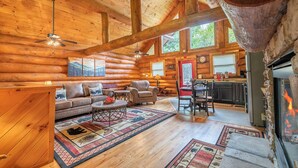 A log cabin interior with a living room, dining area, and kitchen, featuring wooden walls, large windows, and a fireplace.