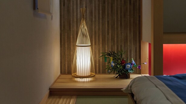 Una habitación con suelo de madera, una cama con colcha azul y gris, una lámpara de pared y una planta pequeña sobre una mesa de madera.