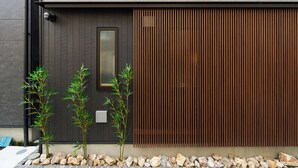Fachada de edificio moderno con un panel vertical oscuro, un ventanal pequeño y tres plantas de bambú.