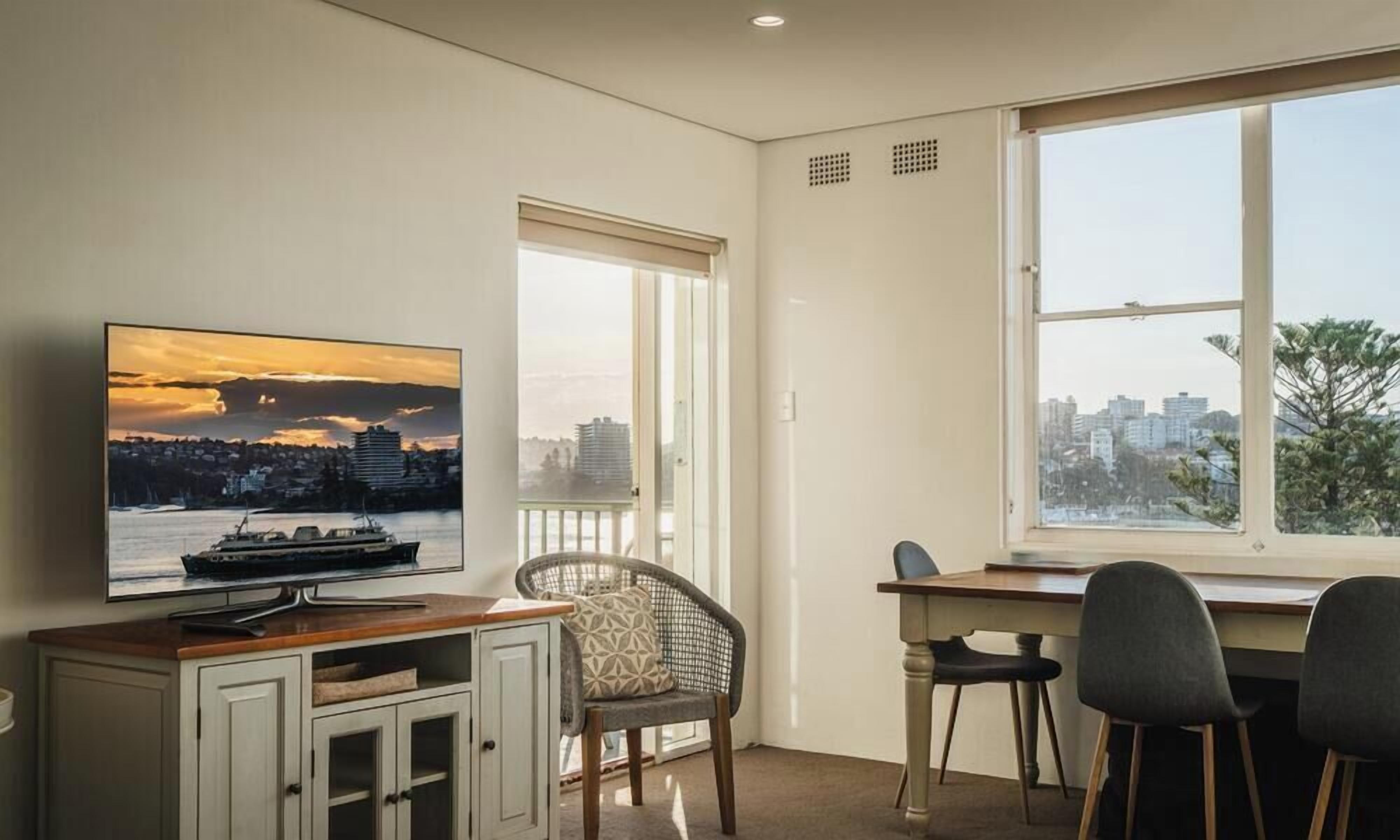 Una habitación con televisión mostrando un paisaje urbano, un escritorio con sillas y una ventana con vista a la ciudad.