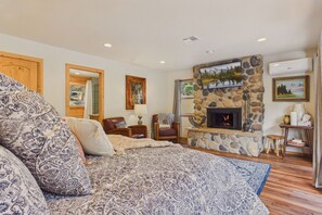 A bedroom with a large bed, a stone fireplace, a TV, and a window with a view. - Beautiful log cabin high above the Rio Ruidoso River. The Log & Order Cabin. (Ruidoso)