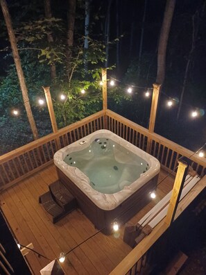Un bain à remous sur une terrasse en bois, illuminé de guirlandes lumineuses, et entouré d’arbres.
