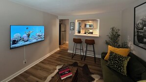 Una sala de estar moderna con una televisión de pantalla plana, una mesa de centro de madera, un sofá verde con un cojín estampado de leopardo, y una zona de cocina con un microondas y dos taburetes de bar.