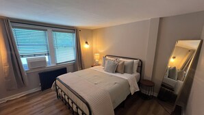 Un dormitorio con cama, lámpara de noche, ventana con persianas, radiador y un espejo en la pared.