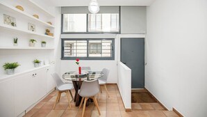 Un coin repas compact, avec une table en verre, quatre chaises et une seule rose rouge. Des étagères ornées de petites plantes et d’objets décoratifs longent les murs.