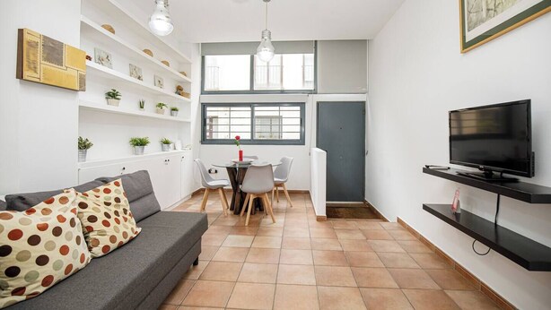 Un salon moderne avec un canapé gris, une table à manger avec des chaises, un téléviseur posé sur un meuble noir et une bibliothèque agrémentée de plantes.