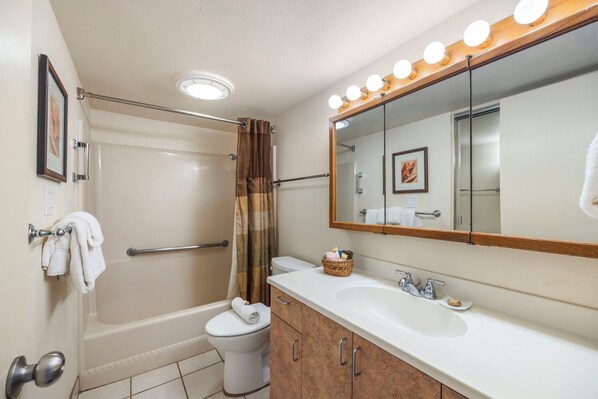 A bathroom with a bathtub, toilet, and sink. There is a shower curtain, a towel rack, and a mirror with lights above the sink. - Kuleana Club - 504 (Lahaina)