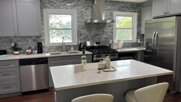 Uma cozinha moderna com armários cinza, uma ilha central com dois banquetes, uma geladeira de aço inoxidável e um fogão a gás.