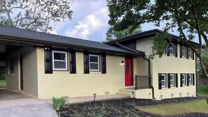 Uma casa de dois andares com porta vermelha, um pequeno alpendre e um jardim frontal bem cuidado.