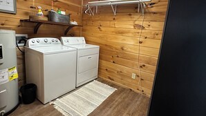 Een wasserette met een wasmachine en droger, een klein schap met schoonmaakmiddelen, een kalender en een zwarte deur.