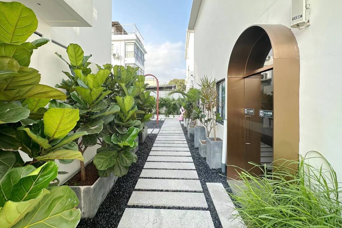 A narrow pathway flanked by potted plants and a building entrance with a sign.