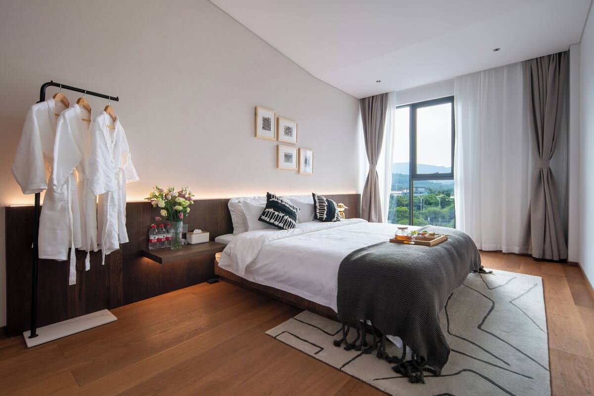 A modern bedroom with a large bed, a bedside table with a vase of flowers, and a view of the outdoors through a large window.