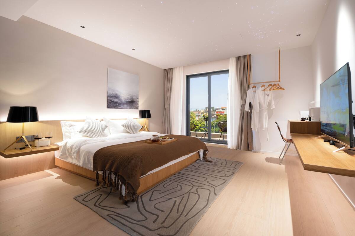 A modern bedroom with a large bed, bedside tables, a TV, and a view of the city through the window.