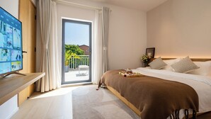 Habitación de hotel con cama, televisor en un mueble, balcón con cortinas y vista a zonas verdes.
