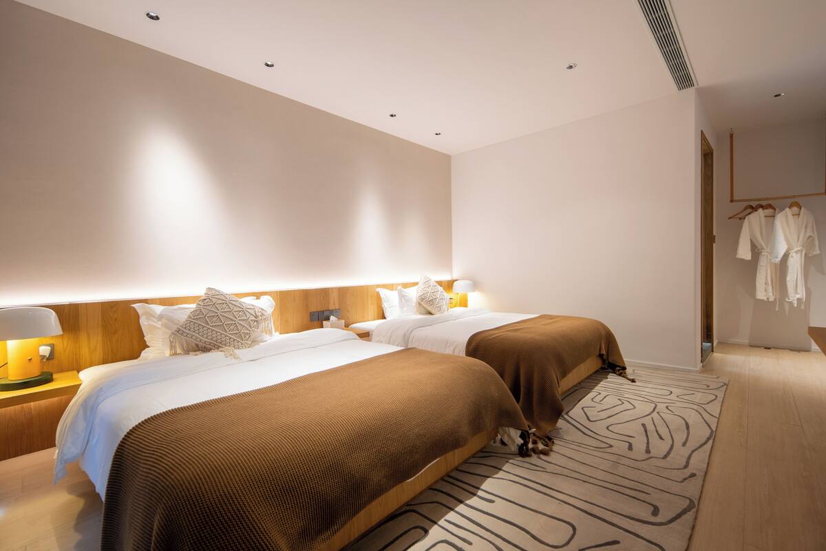 A modern hotel room with two beds, a wooden headboard, a bedside table with a lamp, and a patterned rug.