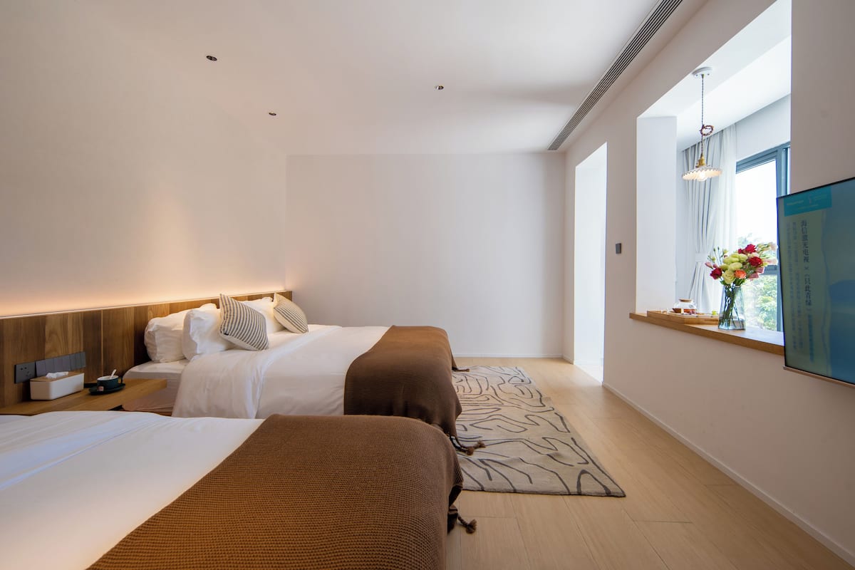 A modern hotel room with two beds, a large window with a view, and a flat-screen TV mounted on the wall.