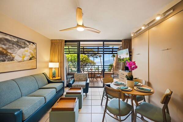 A modern living room with a sofa, a dining table set for four, and a balcony with a view of the ocean. - Kuleana Club - 509 (Lahaina)