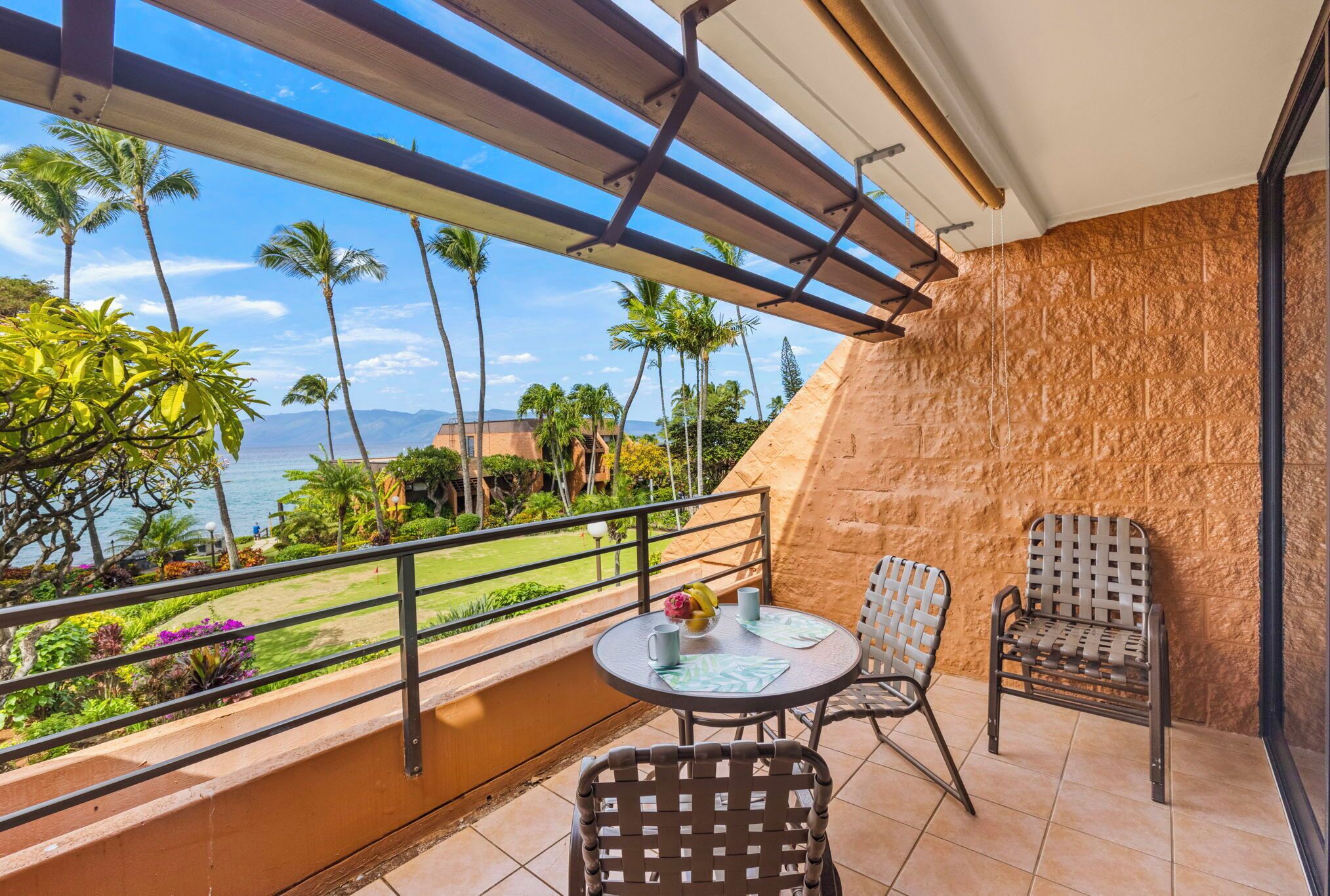 A balcony with a view of the ocean, palm trees, and a stone wall. There are two wicker chairs and a table with a vase of flowers.