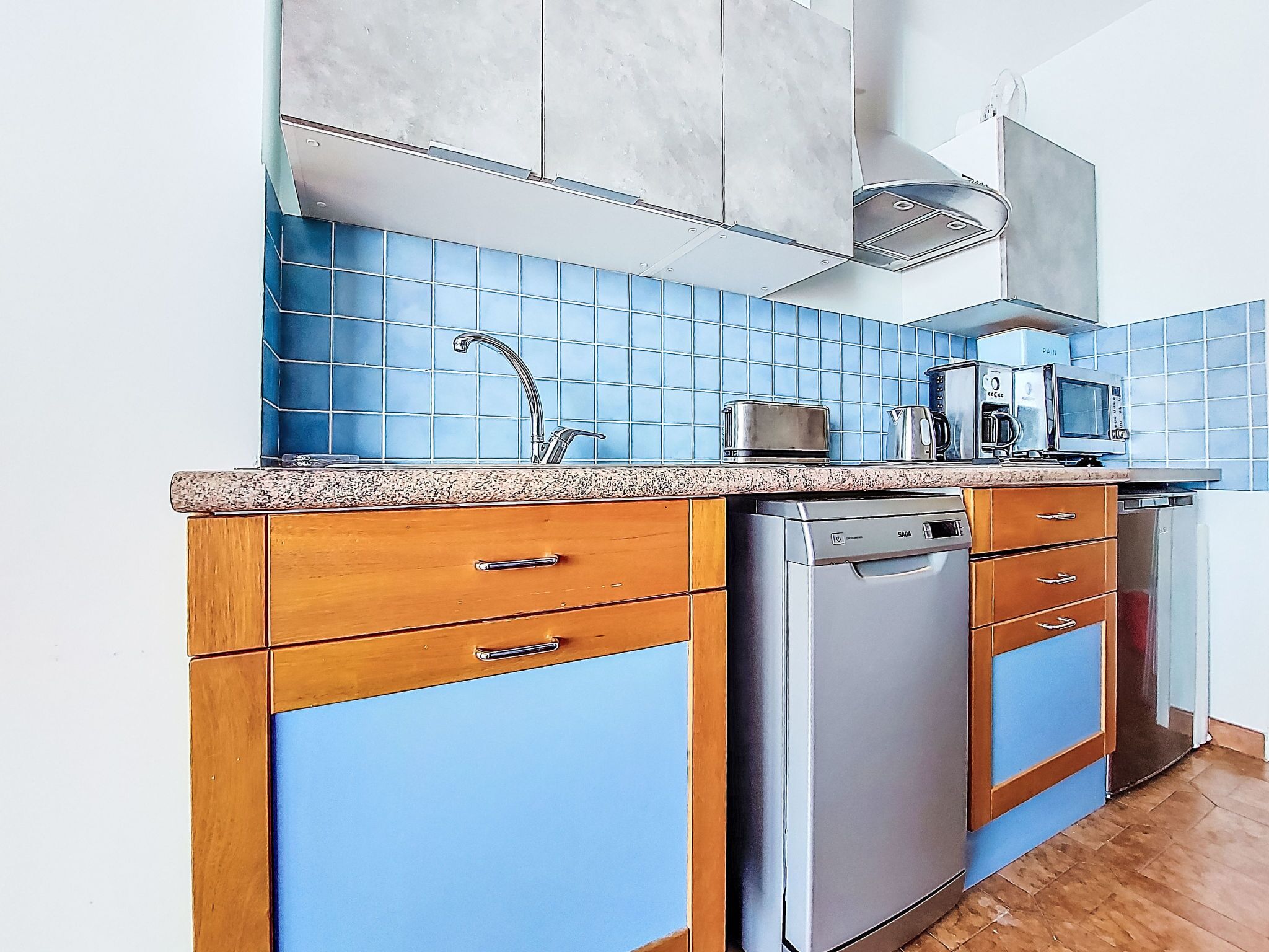 A kitchen with blue tiles, wooden cabinets, a stainless steel dishwasher, and a coffee maker.