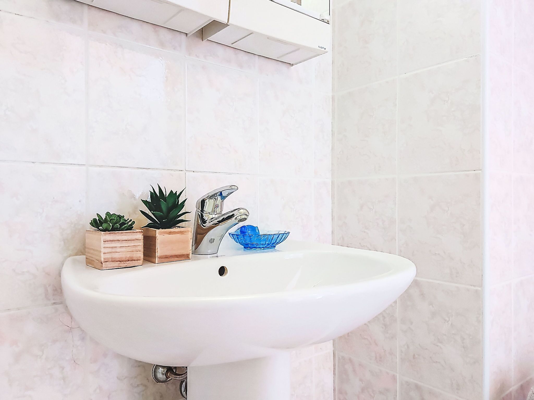 Un lavabo blanc dans une salle de bain, doté d’un robinet moderne, avec deux plantes en pot et un bol en verre bleu posé sur le comptoir.