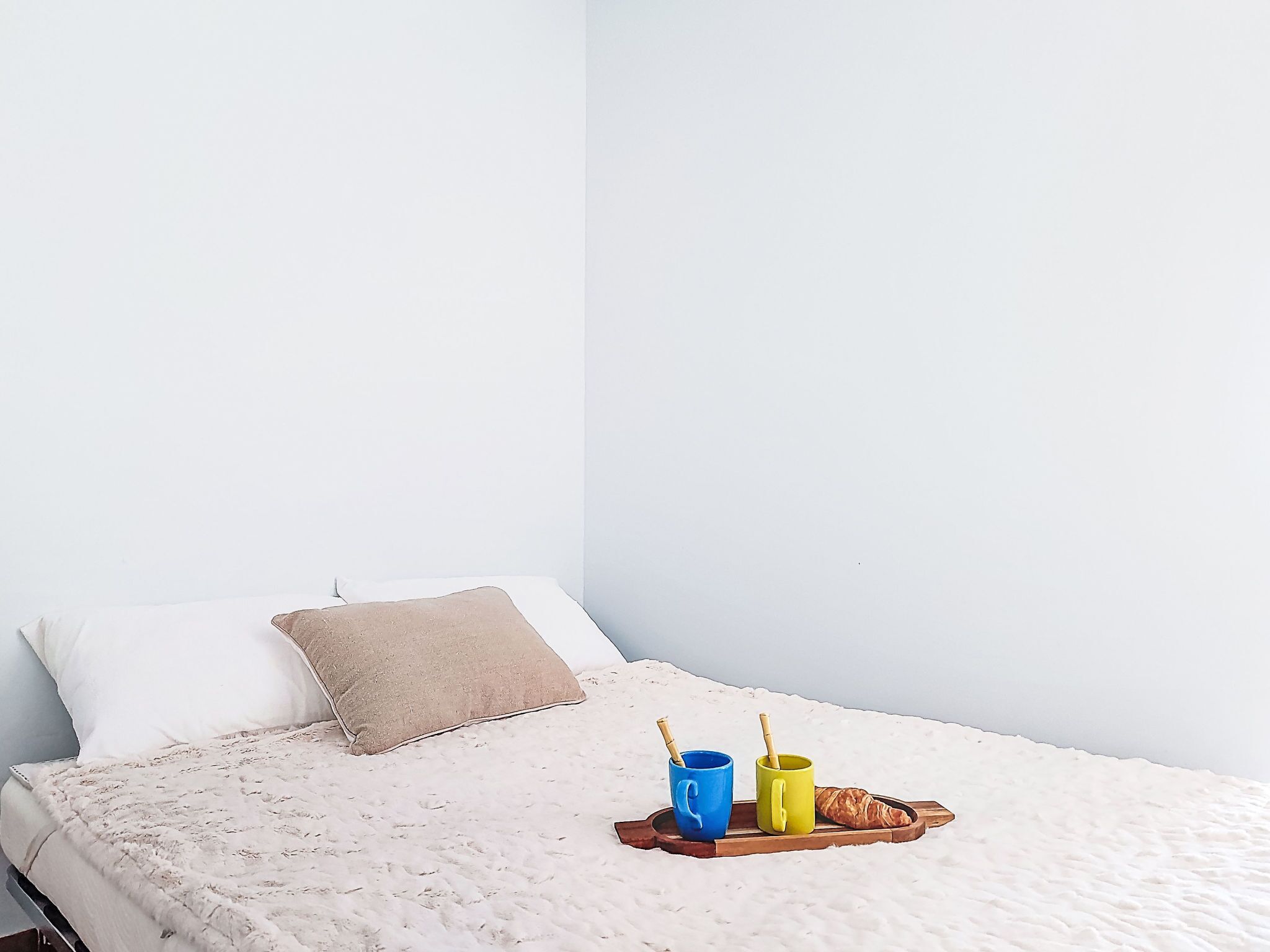 A bed with a white bedspread, a beige pillow, and a tray with two mugs and a croissant.
