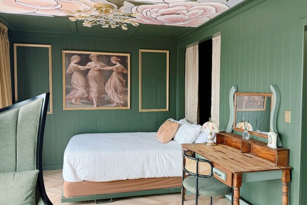 A room with a bed, a desk, and a chair. There are framed pictures on the wall and a chandelier.