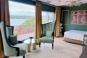 A room with a bed, two armchairs, a small table, and a view of a lake through large windows.