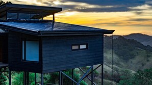 A modern house with a flat roof and large windows, elevated on stilts, overlooking a hilly landscape during sunset. - Stunning view house in Monteverde and reforestation project. (Monteverde)