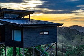 Een modern huis met plat dak en grote ramen, verhoogd op palen, met uitzicht op een glooiend landschap tijdens de zonsondergang.