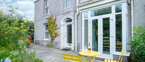 A two-story house with a patio area featuring a yellow table and chairs, a bottle of wine, and a variety of plants and flowers.