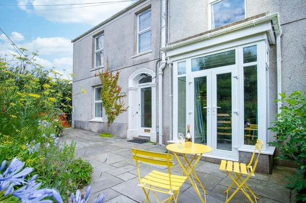 A two-story house with a patio area featuring a yellow table and chairs, a bottle of wine, and a variety of plants and flowers.