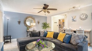 A modern living room with a sectional sofa, a glass coffee table, a ceiling fan, and a dining area in the background.