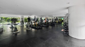 Een sportschool met diverse fitnessapparaten, een grote pilaar en uitzicht op groen buiten.