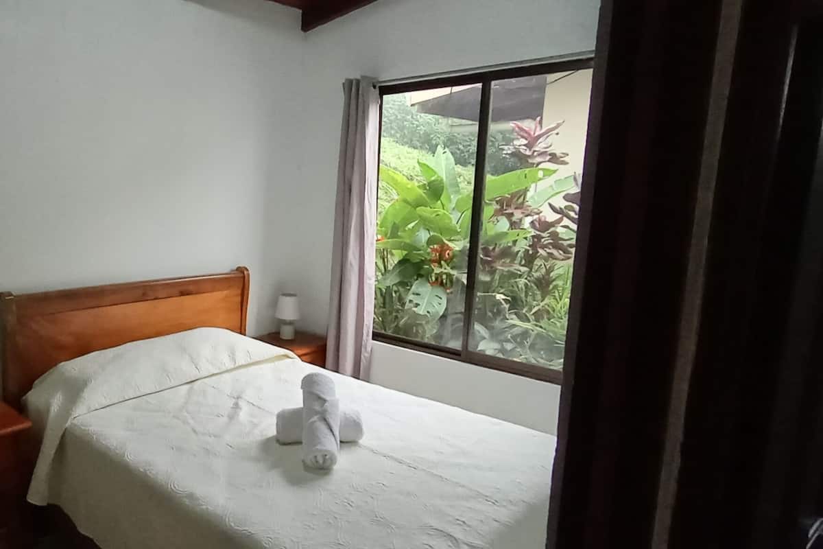 A bedroom with a wooden headboard, a bed with a white bedspread, a bedside table with a lamp, a wall-mounted fan, and a window with a view of greenery.