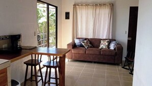 A living room with a brown sofa, two bar stools, a wooden table, and a window with curtains. - Lindora Ranch (Monteverde)