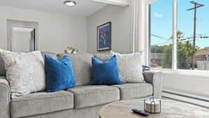 A grey sofa with floral and blue cushions, a round wooden coffee table with a remote control, and a framed painting on the wall.