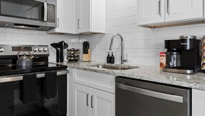 A modern kitchen with white cabinets, a stainless steel microwave, a kettle, a coffee maker, and a dishwasher.