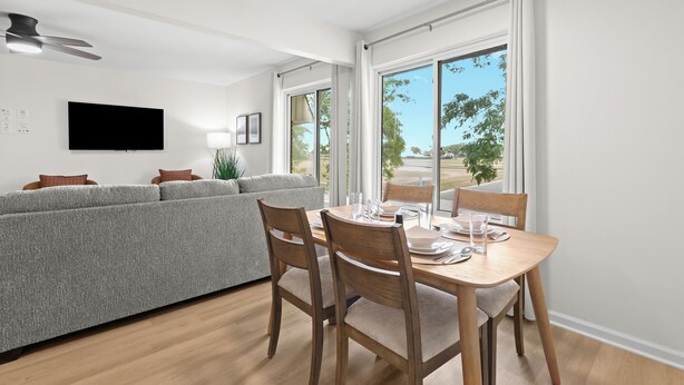 A modern living room with a grey sofa, wooden dining table, and chairs. A flat-screen TV is mounted on the wall, and there is a large window with curtains.
