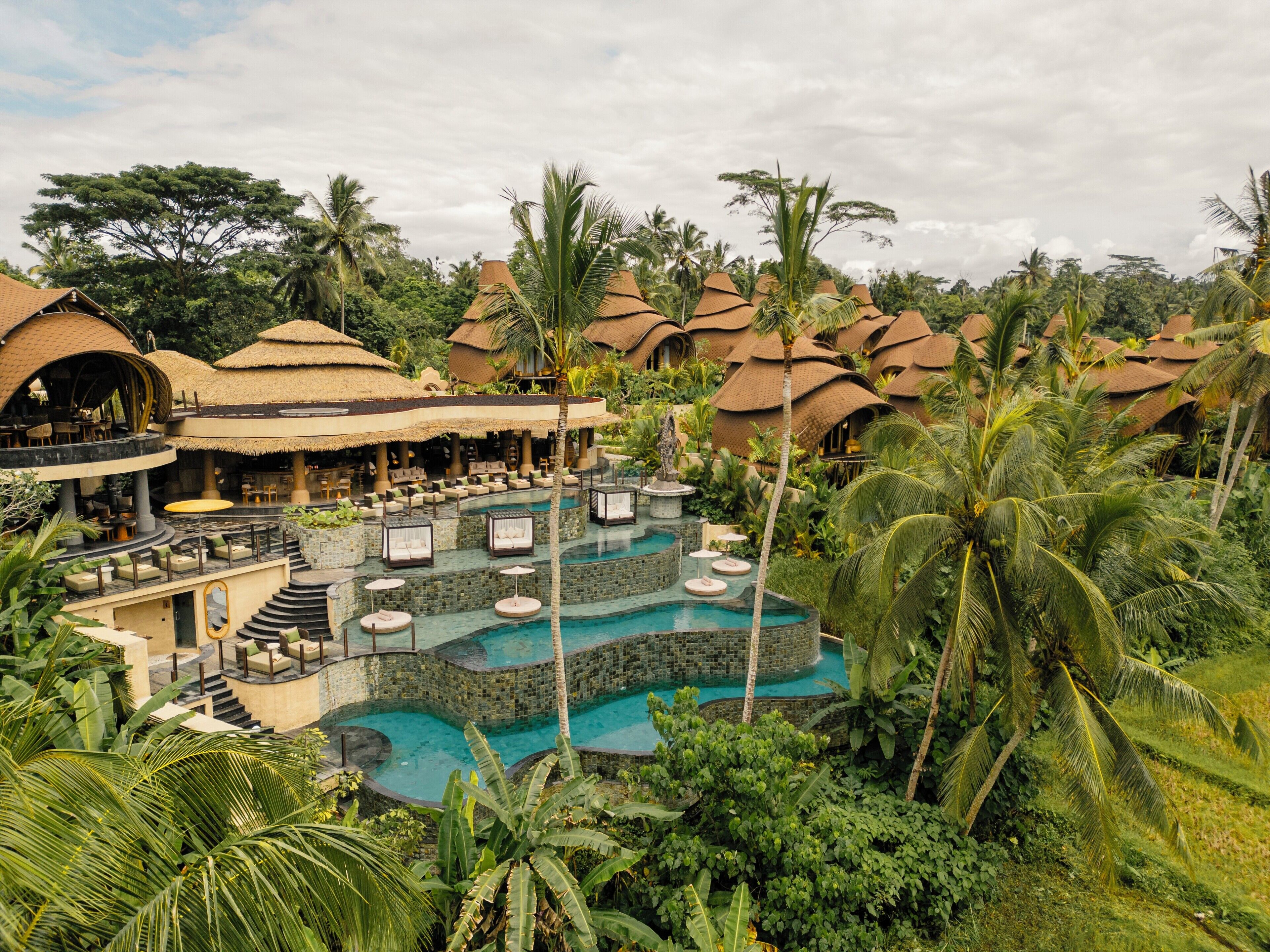 A resort with multiple thatched-roof structures, a central pool area, and lush tropical vegetation.