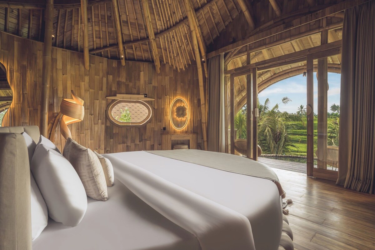 A bedroom with a bed, pillows, a lamp, a wall clock, and a view of greenery outside.