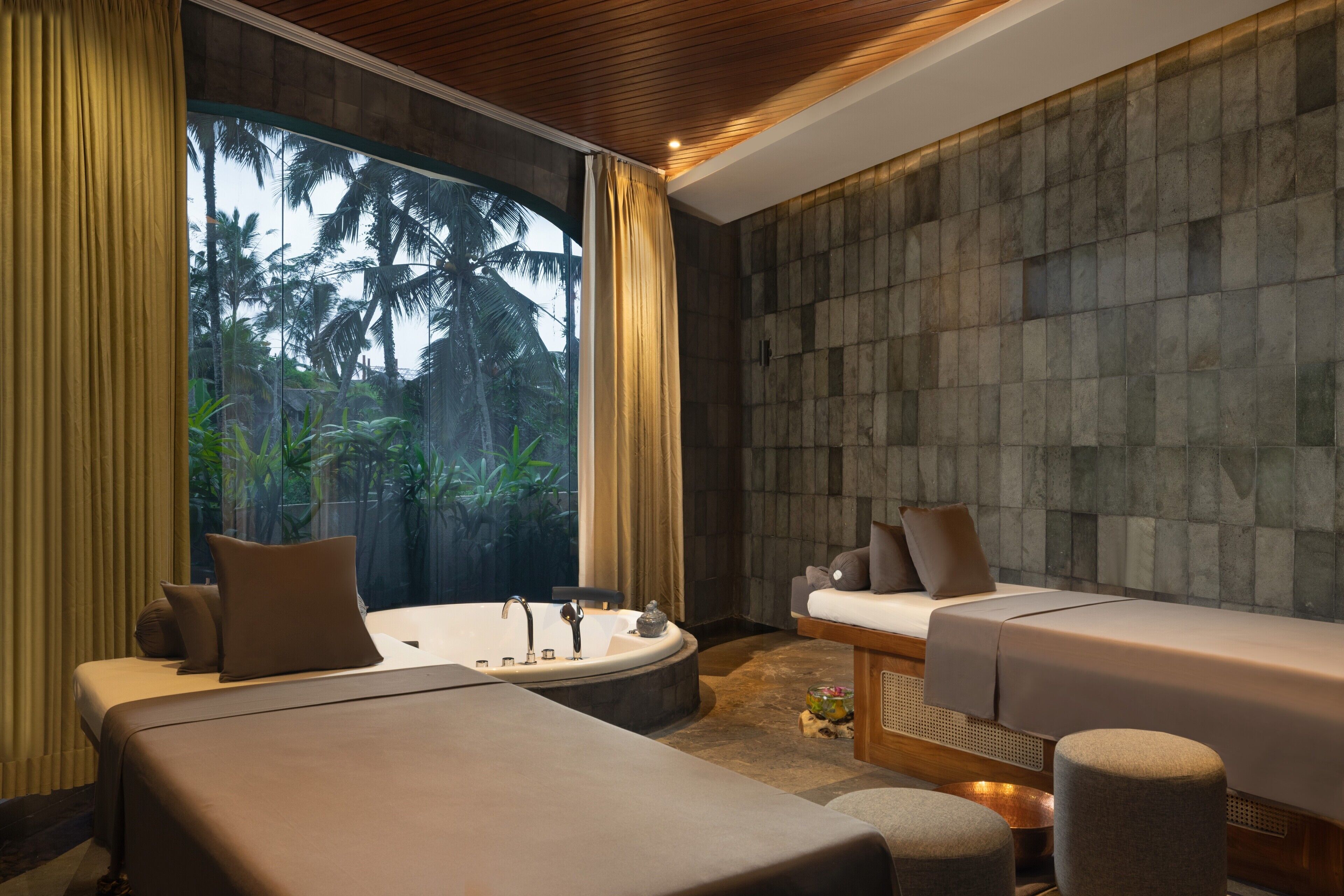 A modern spa room with a large window, a bathtub, a massage table, and a seating area.