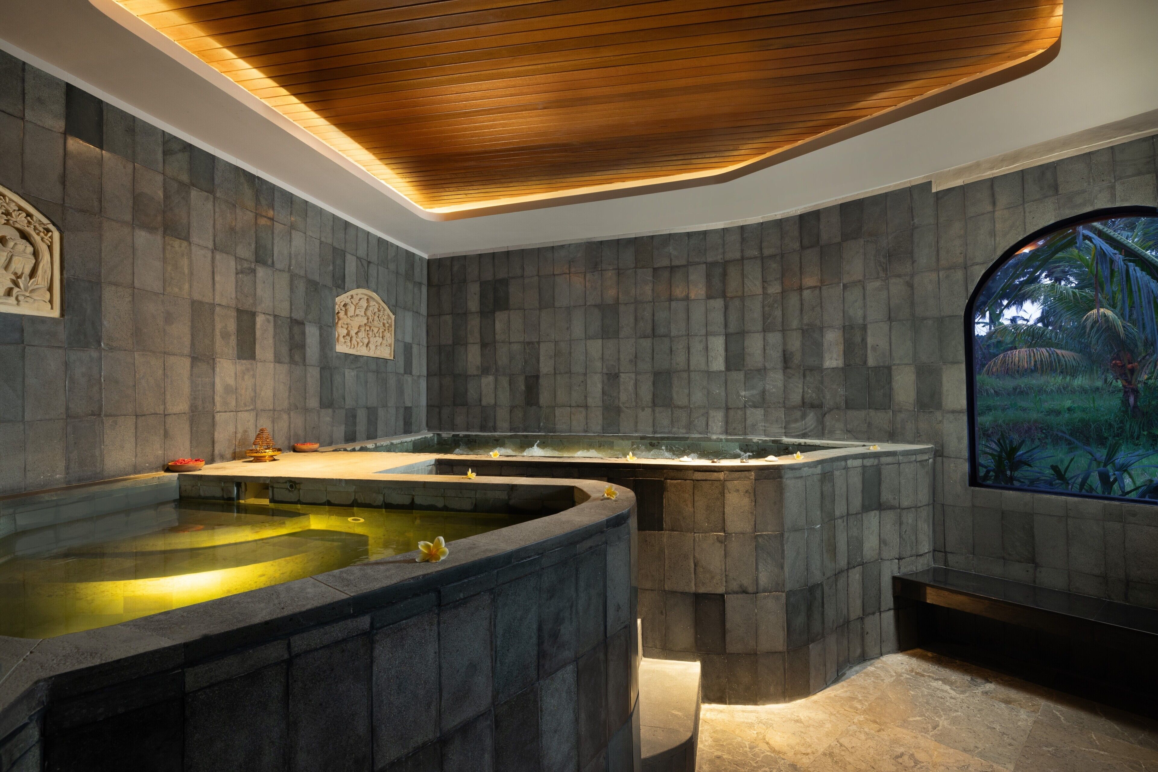 A spa area with a large stone bathtub, a smaller hot tub, and a wall-mounted screen displaying a tropical scene.