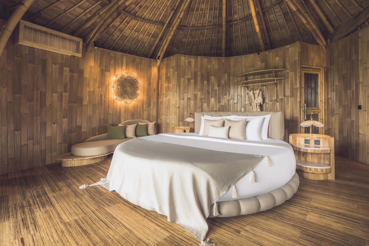 A room with a circular bed, wooden walls, a ceiling fan, and a small table with a lamp.