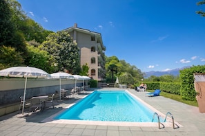 A swimming pool with lounge chairs and umbrellas, surrounded by greenery and a building with multiple balconies.