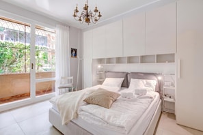 A modern bedroom with a large bed, white walls, and a view of greenery outside.