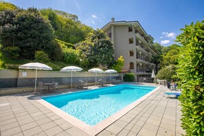 A swimming pool with lounge chairs and umbrellas, surrounded by greenery and a multi-story building.