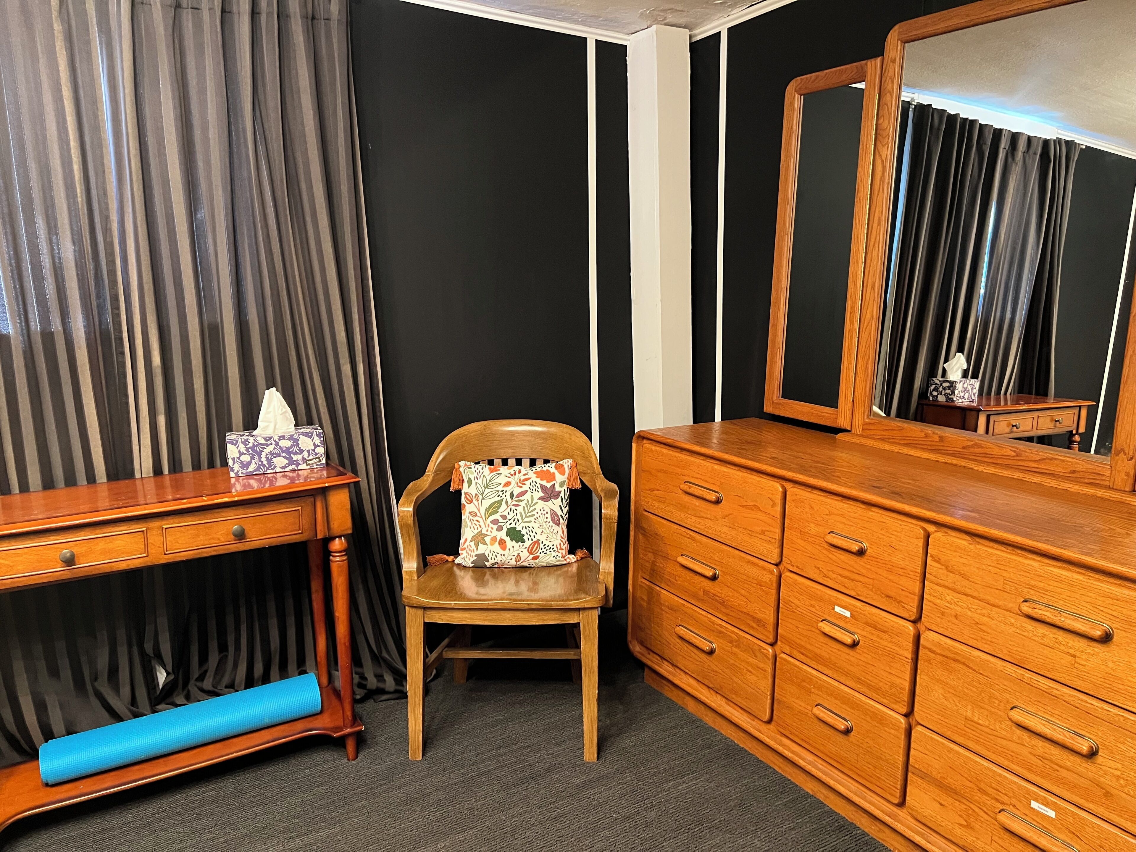 A room with a wooden dresser, a chair with a floral cushion, a small table with a tissue box, and a mirror reflecting a curtain.