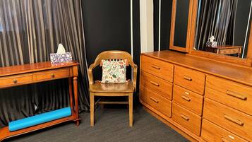 A room with a wooden dresser, a chair with a floral cushion, a small table with a tissue box, and a mirror reflecting a curtain.