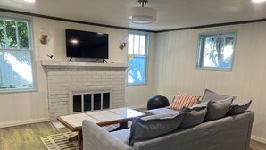 A living room with a grey sofa, a flat-screen TV mounted above a white fireplace, and a window with a view of trees. - Serene Neighborhood Near beach; Under 10 min. to hospitals, universities, & DT (Tacoma)